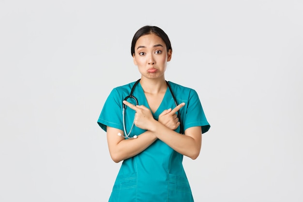 Covid19 Healthcare Workers Pandemiekonzept Unentschlossene und ahnungslose asiatische Krankenschwesternpraktikantin wissen nicht, was sie wählen, wenn sie mit dem Finger zur Seite zeigen und ahnungslosen weißen Hintergrund zucken