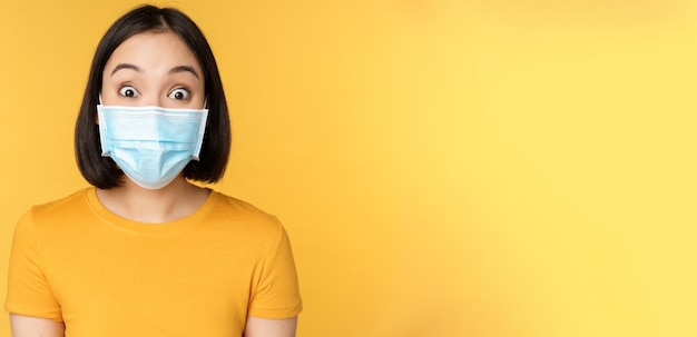 Covid19 und medizinisches Konzept Nahaufnahme des Porträts einer asiatischen Frau in Gesichtsmaske, die überrascht und erstaunt über Nachrichten auf gelbem Hintergrund aussieht