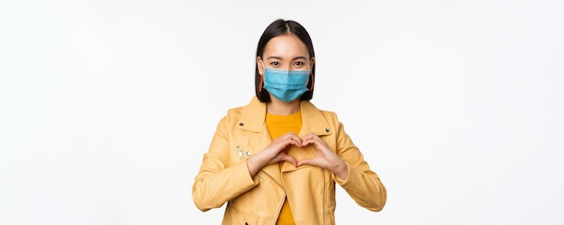 Covid19 und Personenkonzept Junge asiatische Frau in medizinischer Gesichtsmaske, die Herzgeste zeigt und mit sorgfältig weißem Hintergrund lächelt