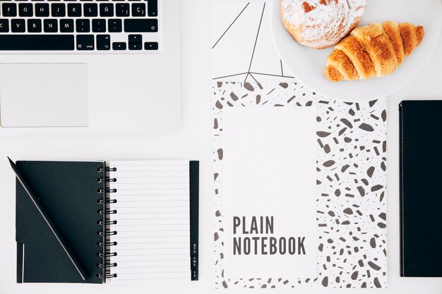 Croissant; einfaches Notizbuch; Laptop; Bleistift und Spiralblock auf weißem Schreibtisch