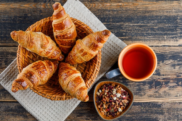 Croissant im Korb mit Tasse Tee, getrocknete Kräuter flach auf Holz- und Küchentuch legen