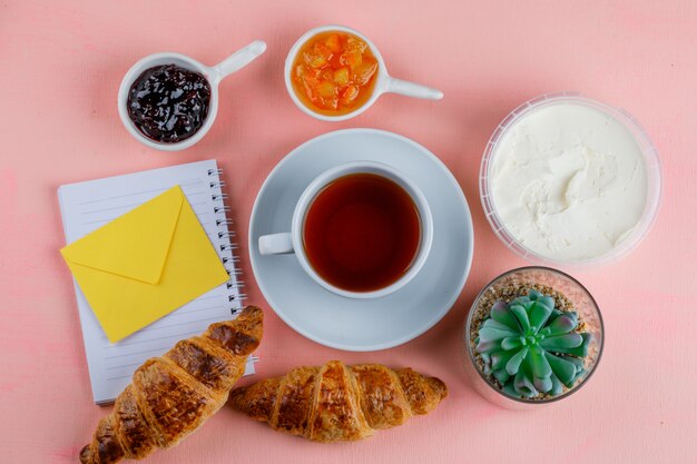Croissant mit Frischkäse, Tee, Marmelade, Pflanze, Umschlag, Notizbuch auf rosa Tisch, flach liegen.