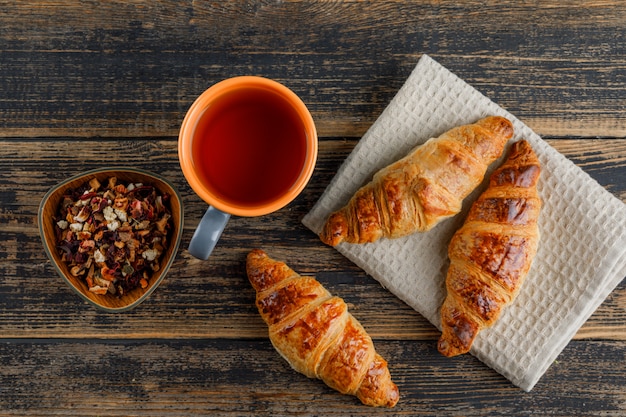 Croissant mit Tee in einer Tasse, getrocknete Kräuter flach auf Holz- und Küchentuch gelegt