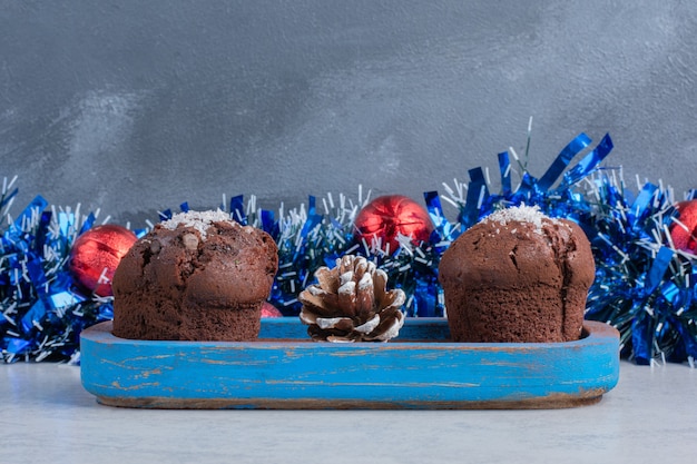 Cupcakes und ein Tannenzapfen auf einer Platte neben einer Girlande und Kugeln auf Marmoroberfläche