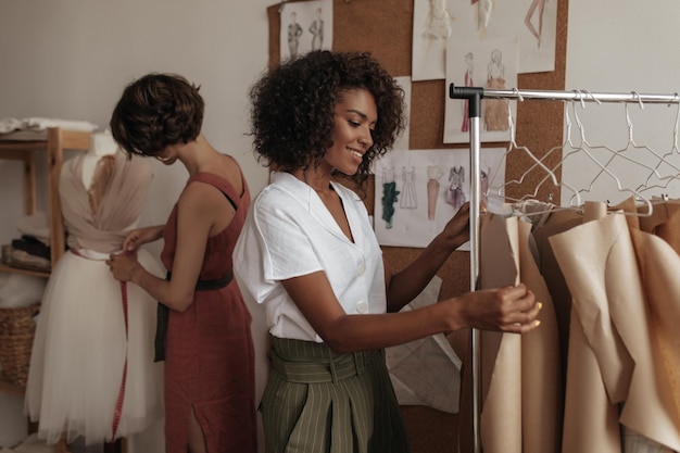 Curly attraktive dunkelhäutige Frau in weißer Bluse und Khaki-Shorts arbeitet als Modedesignerin Kurzhaarige Dame entwirft Kleid