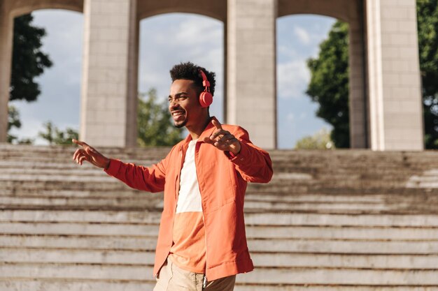 Curly Brunet dunkelhäutiger Mann in orangefarbener Jacke und stylischem T-Shirt tanzt und hört draußen Musik in roten Kopfhörern