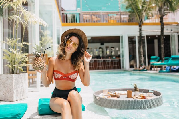 Curly gewinnende Frau in der Sonnenbrille, die Cocktail im Resort genießt. Außenaufnahme der herrlichen gebräunten Frau mit Ananas, die in der Nähe des Pools am sonnigen Tag sitzt.
