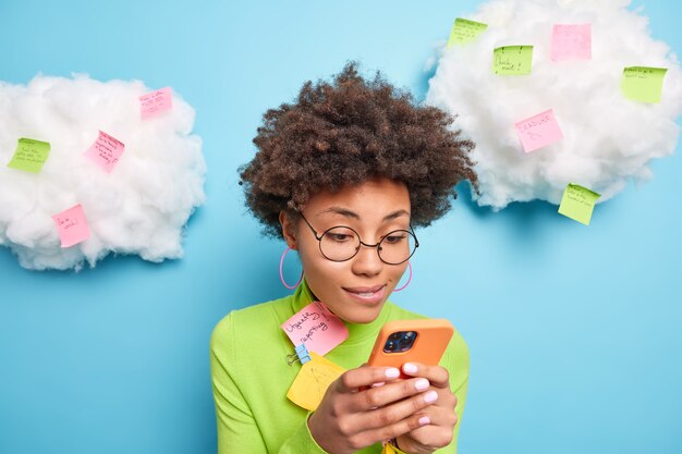 Curly haired ethnische Frau konzentriert auf Smartphone-Display sendet Textnachrichten trägt runde Brille Rollkragenpullover umgeben von Aufklebern mit schriftlichen Ideen und Plänen