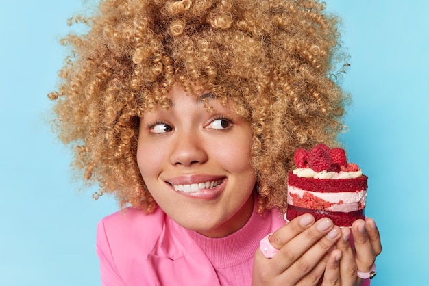 Curly haired schöne frau beißt lippen sieht appetitanregend stück kuchen mit himbeeren hat naschkatzen fühlt versuchung, köstliches dessert isoliert über blauem hintergrund zu essen zuckersucht