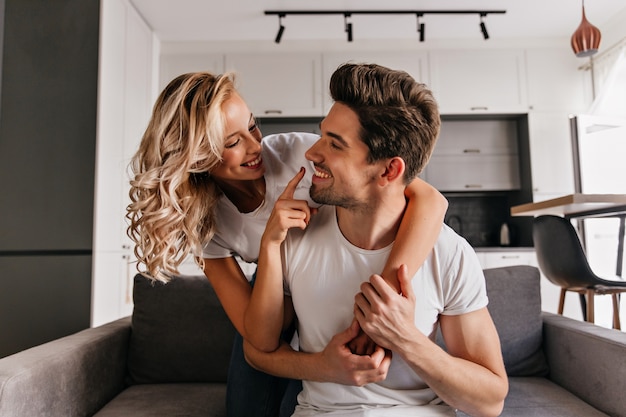 Curly romantisches Mädchen, das die Nase des Freundes berührt. Innenporträt des fröhlichen Paares, das zusammen im Wohnzimmer aufwirft.