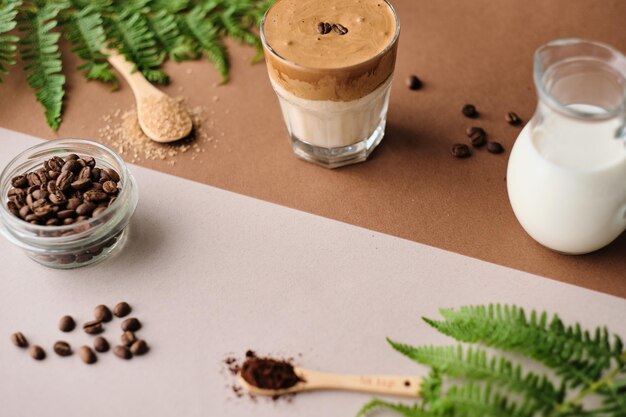 Dalgona-Eiskaffee mit Mandelmilch und braunen Zuckerkaffeebohnen und grüner Pflanze auf einem beigen Blatt Ein Glas trendiges, flauschiges koreanisches Getränk mit Schlagsahne xAxA