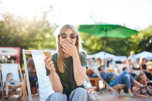 Dame, die Eis lachend isst. Porträt der jungen Frau, die in einem Park an einem sonnigen Tag sitzt, der Eis isst