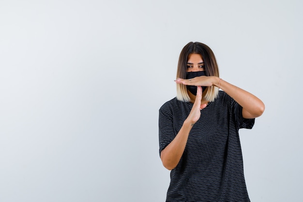 Dame, die zeitpausengeste im schwarzen kleid, in der medizinischen maske zeigt und entschlossen aussieht. vorderansicht.