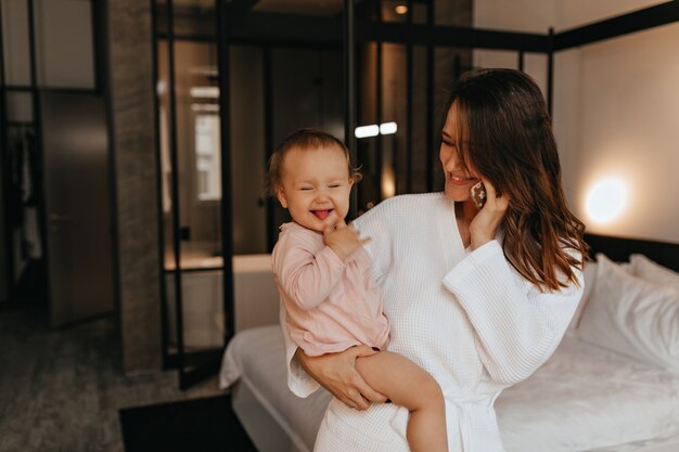 Dame im Bademantel in ausgezeichneter Stimmung spricht am Telefon und sitzt auf dem Bett mit frechen weiblichem Baby, das ihre Zunge zeigt.