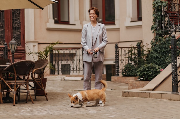 Dame im grauen Anzug geht mit ihrem Corgi. Porträt der glücklichen Frau in der Jacke und in der Hose, die mit Hund außerhalb aufwerfen