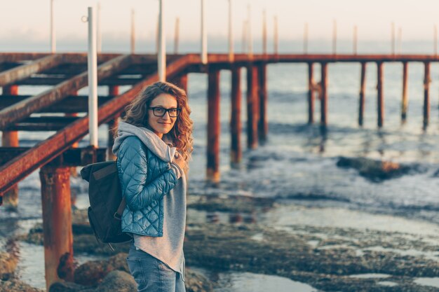 Dame im grauen Kapuzenpulli und im blauen Mantel stehend und lächelnd im Strand während des Sonnenuntergangs