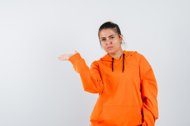 Dame im orangefarbenen Hoodie zeigt eine einladende Geste und sieht vernünftig aus looking