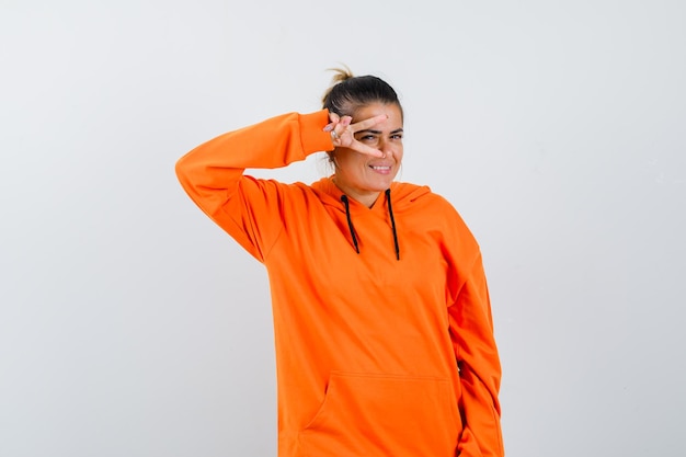 Dame im orangefarbenen Hoodie zeigt V-Zeichen auf dem Auge und sieht glücklich aus