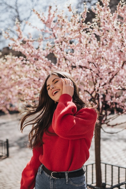 Dame im roten Pullover lacht und spielt mit Haaren vor dem Hintergrund von Sakuras. Glückliche Frau in der Kaschmir-Strickjacke und in den Jeans, die draußen aufwerfen