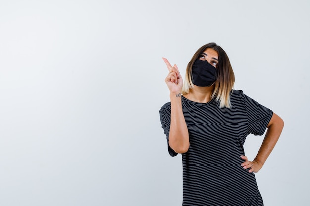 Dame im schwarzen Kleid, medizinische Maske zeigt auf obere linke Ecke und sieht nachdenklich aus, Vorderansicht.