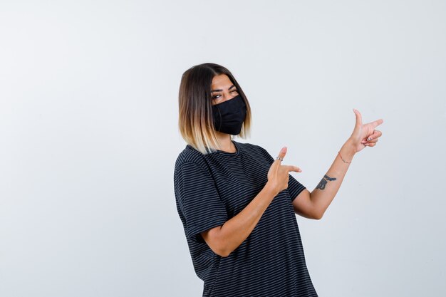 Dame im schwarzen Kleid, medizinische Maske zeigt nach rechts und sieht selbstbewusst aus, Vorderansicht.