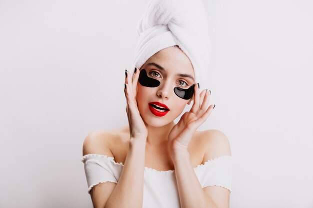 Dame im weißen Handtuch auf ihrem Kopf posiert mit schwarzen Flecken für das Auge. Mädchen mit rotem Lippenstift liebt morgendliche Spa-Behandlungen.