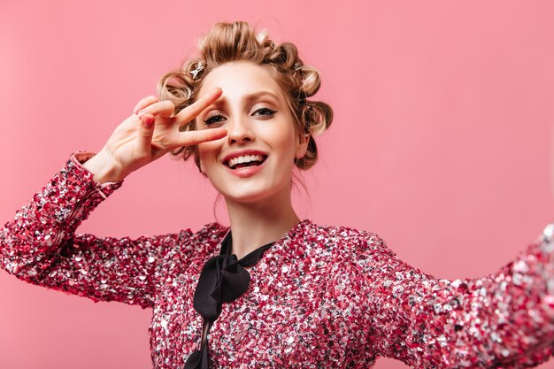 Dame in Bluse mit Pailletten nimmt Selfie und zeigt Friedenszeichen auf rosa Wand