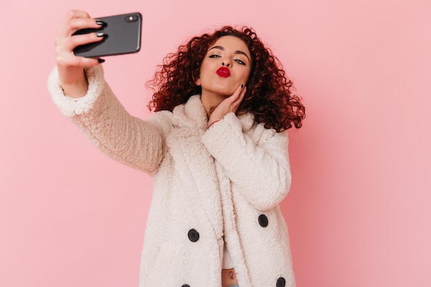 Dame in weißem Mantel und kurz geschnittenem Oberteil bläst Kuss und macht Selfie Porträt einer lockigen Dame mit hellen Lippen auf rosa Hintergrund