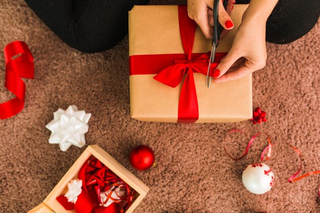 Dame mit Geschenkbox und Schere in der Nähe von dekorativen Bögen, Bällen und Band auf Teppich