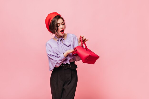 Kostenloses Foto dame mit roten lippen, die in tasche schauen. porträt des mädchens im hut und im eleganten outfit auf rosa hintergrund.