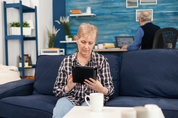 Dame mittleren Alters, die sich auf dem Sofa entspannt und auf einem Tablet-PC liest