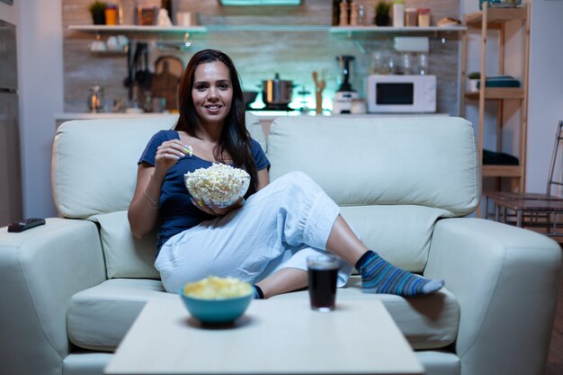 Dame ruht sich mit Snacks und Saft aus und sieht sich nachts einen Film an, der auf einem bequemen Sofa im offenen Wohnzimmer sitzt. Aufgeregt, amüsiert allein zu Hause, entspannen Sie beim Fernsehen und wechseln Sie die Kanäle mit Fernbedienung