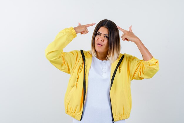 Dame zeigt auf ihren Kopf in T-Shirt, Jacke und sieht verwirrt aus. Vorderansicht.