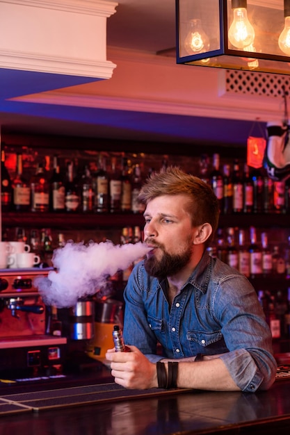 Dampfen. Verdampfender Mann in einer Dampfwolke. Das Foto wird in einer Vape-Bar aufgenommen. Vape-Shop
