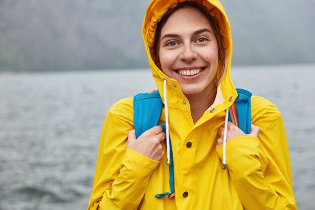 Das abgeschnittene Bild einer optimistischen europäischen Frau trägt eine gelbe Kapuze, trägt einen Rucksack und hat ein breites Lächeln