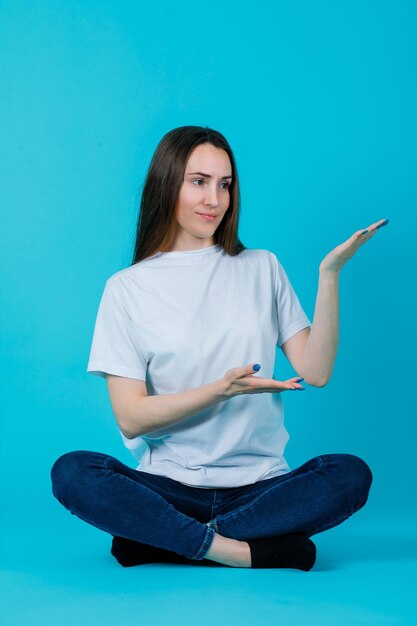 Das auf dem Boden sitzende Mädchen zeigt mit den Händen nach rechts, indem es auf blauem Hintergrund auf dem Boden sitzt