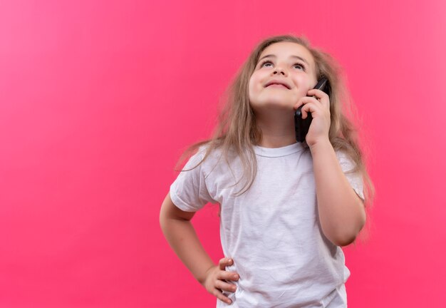 Das Aufschauen des kleinen Schulmädchens, das weißes T-Shirt trägt, spricht am Telefon, legte ihre Hand auf Hüfte auf lokalisiertem rosa Hintergrund