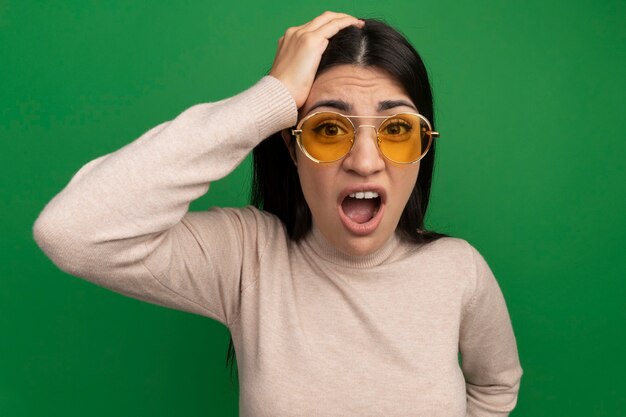 Das besorgte hübsche brünette kaukasische Mädchen in der Sonnenbrille legt Hand auf Kopf und schaut Kamera auf Grün