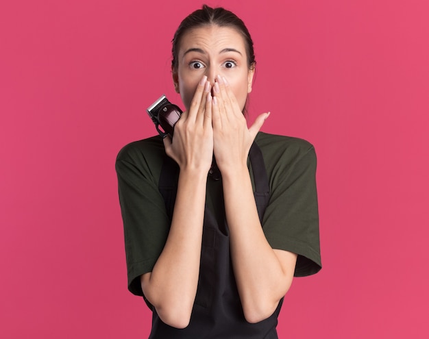 Kostenloses Foto das besorgte junge brünette friseurmädchen in der uniform legt die hände auf den mund und hält die haarschneidemaschine auf rosa