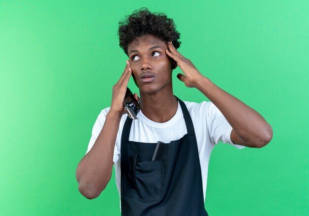 Das Betrachten der Seite beeindruckte den jungen afroamerikanischen männlichen Friseur, der Uniform trägt, die Haarschneidemaschinen hält und Finger auf Stirn lokalisiert auf grünem Hintergrund setzt