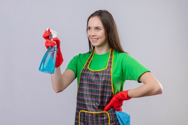 Das Betrachten der Seitenreinigung der jungen Frau, die Uniform in den roten Handschuhen hält, die Reinigungsspray halten, legte ihre Hand auf Hüfte auf isolierte weiße Wand