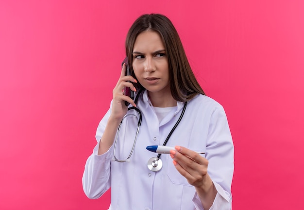 Das Betrachten des jungen Doktormädchens der Seite, das medizinisches Kleid des Stethoskops hält, das Thermometer hält, spricht am Telefon auf lokalisiertem rosa Hintergrund