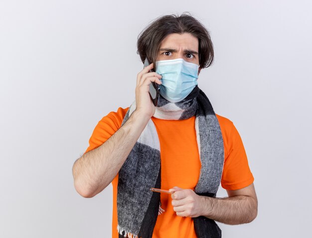 Das Betrachten des jungen kranken Mannes der Kamera, der Schal und medizinische Maske trägt, spricht am Telefon, das Thermometer hält, das auf weißem Hintergrund lokalisiert wird