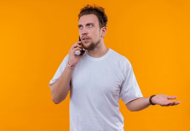 Das Betrachten des jungen Mannes der Seite, der weißes T-Shirt trägt, spricht am Telefon, das Hand auf Seite auf lokalisiertem orange Hintergrund hält