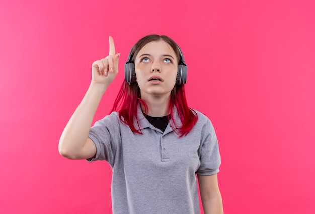 Das Betrachten des jungen schönen Mädchens, das graues T-Shirt im Kopfhörer trägt, zeigt Finger nach oben auf lokalisiertem rosa Hintergrund
