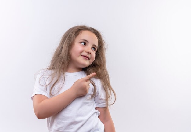 Das Betrachten des kleinen Schulmädchens der Seite, das weißes T-Shirt trägt, zeigt zur Seite auf isolierter weißer Wand