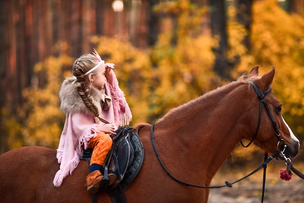 Das bezaubernde kleine Mädchen, das wie eine Prinzessin gekleidet wird, reitet ein Pferd um den Herbstwald