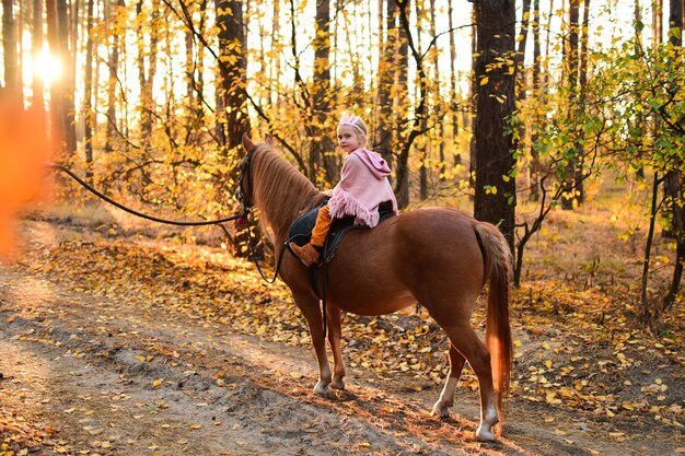 Das bezaubernde kleine Mädchen, das wie eine Prinzessin gekleidet wird, reitet ein Pferd um den Herbstwald