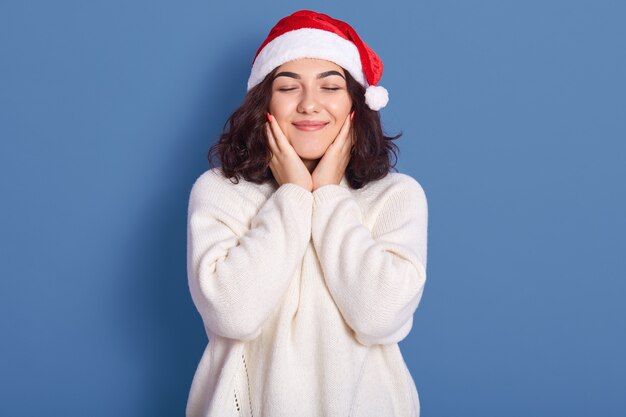 Das Bild der netten jungen Frauen, die warmen weißen Winterpullover und Weihnachten tragen, die mit geschlossenen Augen und Händen auf Wange aufwerfen, isoliert auf blauem Hintergrund posieren, sieht chrming und niedlich aus. Neues Ohrkonzept.