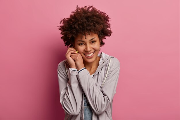 Das Bild des positiven charmanten tausendjährigen Mädchens hält die Hände in der Nähe des Gesichts zusammen, schaut mit zartem Ausdruck, hört etwas Angenehmes, posiert über der rosa Wand. Konzept der positiven Gefühle